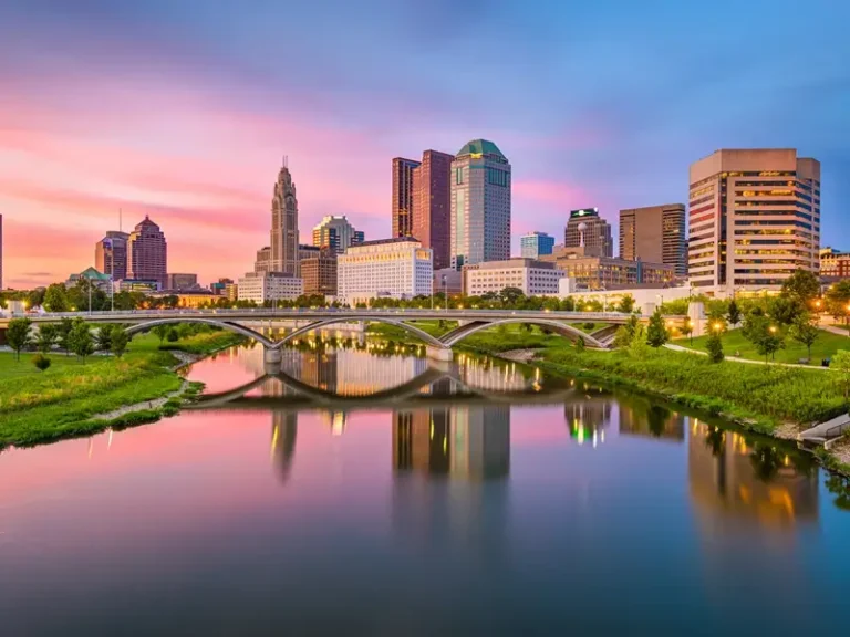 City skyline at sunset reflected in a river beneath a bridge.