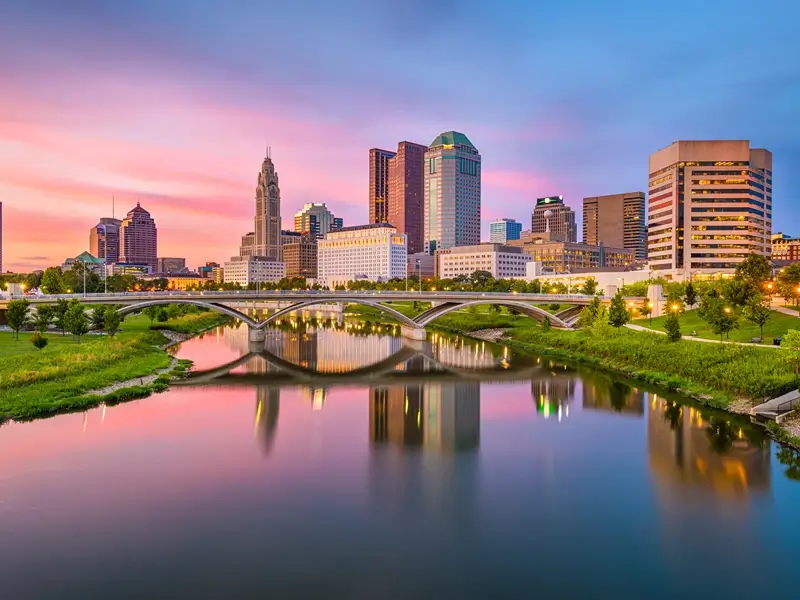 City skyline at sunset reflected in a river beneath a bridge.