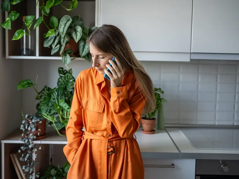 Woman in an orange dress talking on a phone in a kitchen.